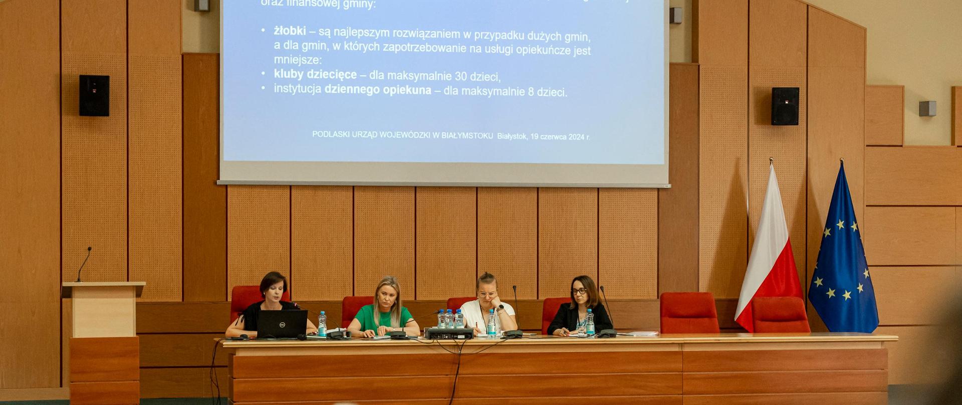 Wyłożona drewnem duża sala konferencyjna, cztery kobiety za długim stołem prezydialnym. Na ścianie za nimi duży ekran z prezentacją. Na pierwszym planie publiczność skierowana ku prelegentkom.