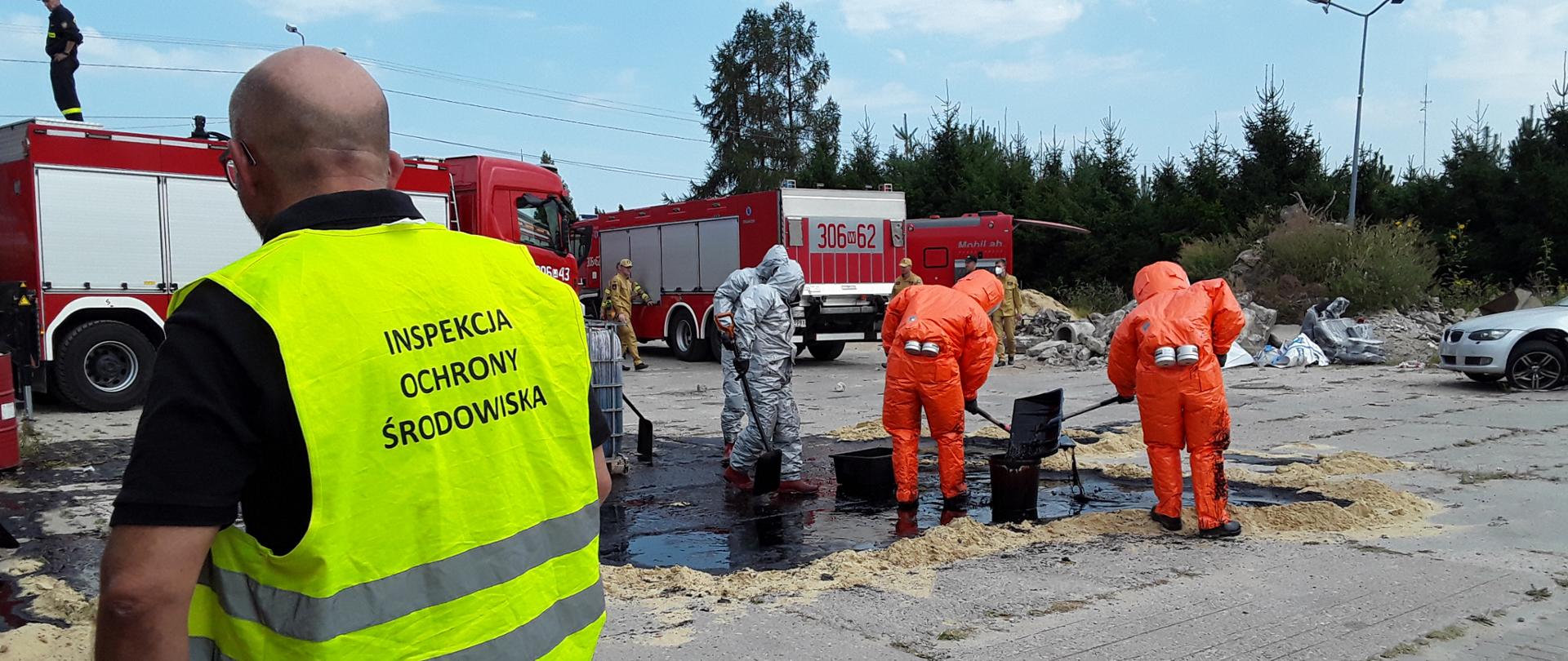 Inspektor Wojewódzkiego Inspektoratu Ochrony Środowiska w Warszawie monitoruje pracę czterech funkcjonariuszy Państwowej Straży Pożarnej. Są oni ubrani w srebrne i pomarańczowe stroje ochronne i na betonowym podłożu utworzyli piaskowy wał w celu zatrzymania rozprzestrzeniania się rozlewiska czarnej substancji. W tle widać dwa wozy bojowe Państwowej Straży Pożarnej.