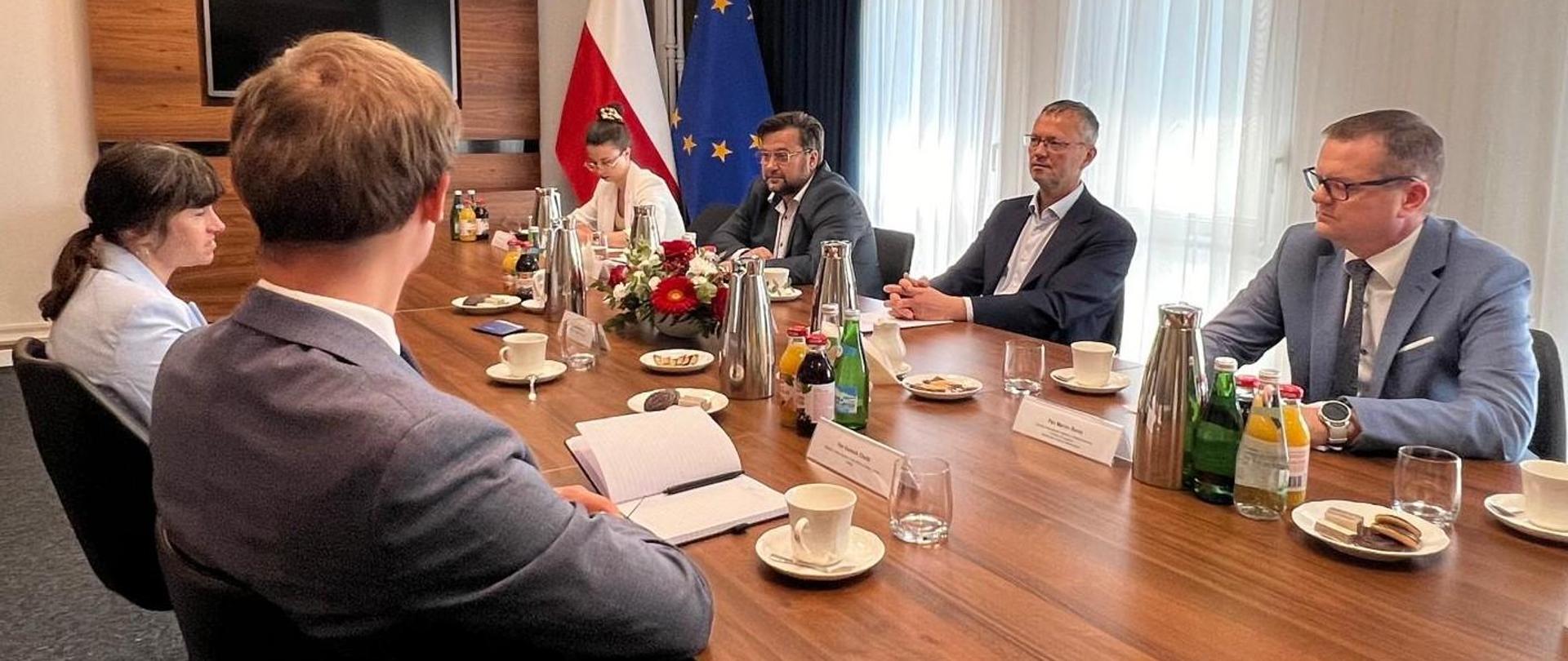 Z lewej strony stołu siedzą przedstawiciele Boeinga, z prawej strony delegacja MAP. Na stole stoją dzbanki, butelki, filiżanki i kwiaty. W tle ekran i flagi - polska i unijna. 