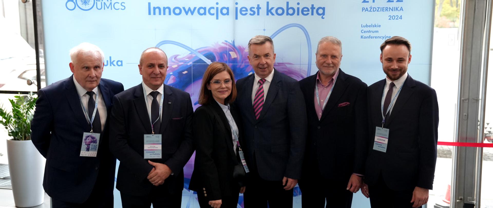 Na tle ścianki z napisem "Innowacja jest kobietą" stoi grupa sześciu osób - pośrodku minister Wieczorek, kobieta w czarnej marynarce i czterech mężczyzn w czarnych garniturach.