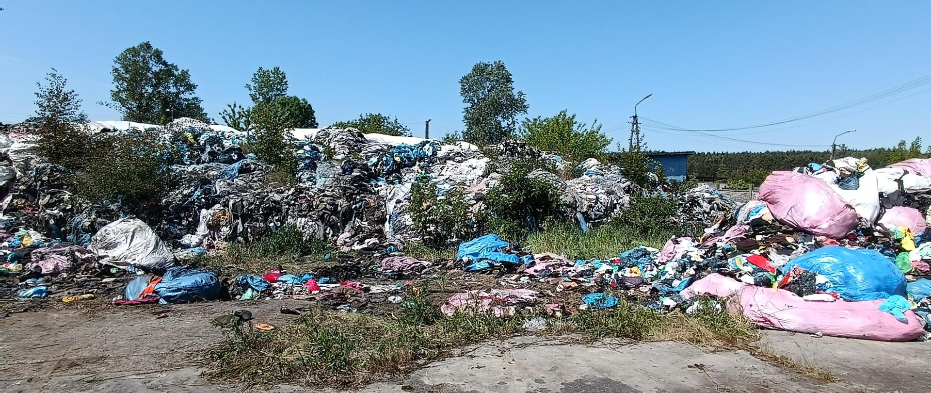 Pryzma częściowo spalonych odpadów komunalnych o powierzchni ok. 600 metrów kwadratowych znajduje się na nieutwardzonym podłożu. Po prawej stronie widać kolorowe torby plastikowe. 