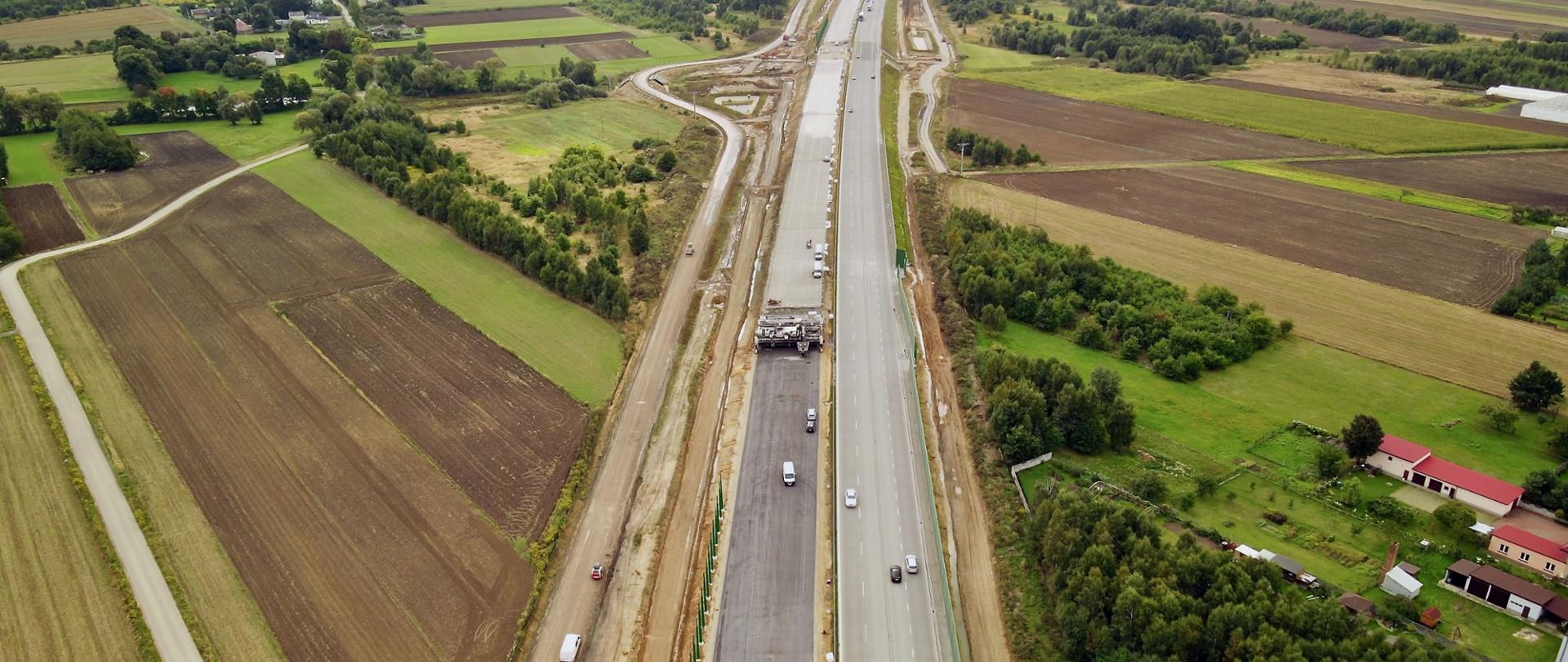 Fotografia z lotu ptaka przedstawiająca budowaną autostradę A1. W centralnej części fotografii znajdują się jezdnie. W prawej części - jezdnia wykonana po której już dwukierunkowo poruszają się pojazdy. Na drugiej jezdni - po lewej stronie - widoczna maszyna do układania nawierzchni betonowej. 
