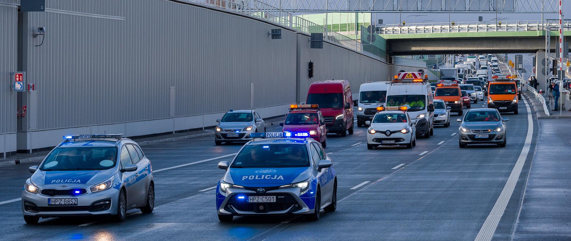 Zdjęcie przedstawia scenę na wielopasmowej drodze z wieloma pojazdami, w tym dwoma radiowozami z migającymi niebieskimi światłami, czerwonym vanem oraz kilkoma innymi samochodami i pojazdami serwisowymi GDDKiA. Wszystkie pojazdy poruszają się za dwoma radiowozami na pierwszym planie.