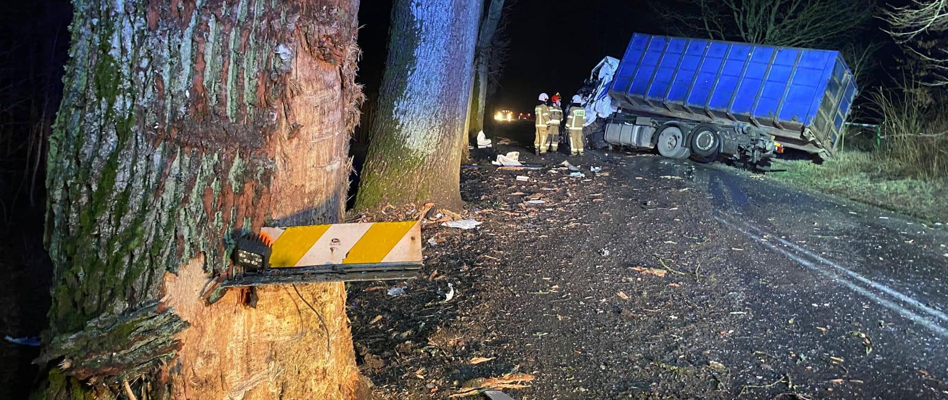 Uszkodzony pojazd ciężarowy stoi w poprzek drogi. Na jezdni znajdują się elementy uszkodzonego pojazdu. Po lewej, wzdłuż jezdni znajdują się drzewa, pierwsze z ich osi ślady uderzenia. w tle widoczne sylwetki strażaków. 