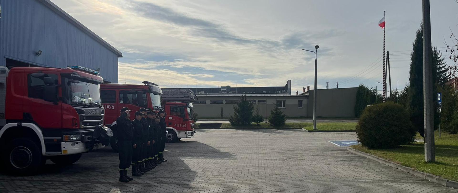 Zdjęcie obrazuje strażaków w umundurowaniu koszarowym. Strażacy znajdują się na tle samochodów ratowniczo-gaśniczych. Po prawej stronie widać maszt, na którym zawieszona jest biało-czerwona flaga.