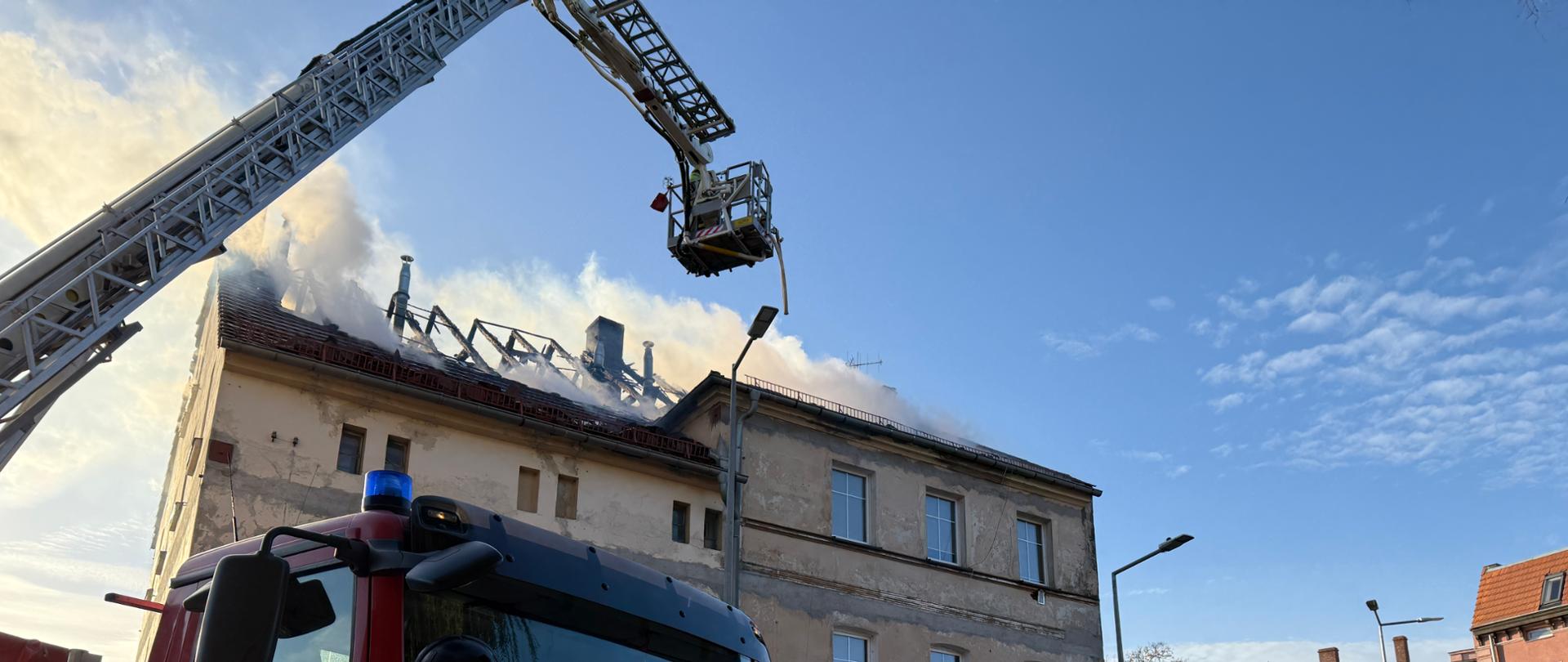 Plac na którym stoi samochód pożarniczy, w tle pali się budynek wielorodzinny