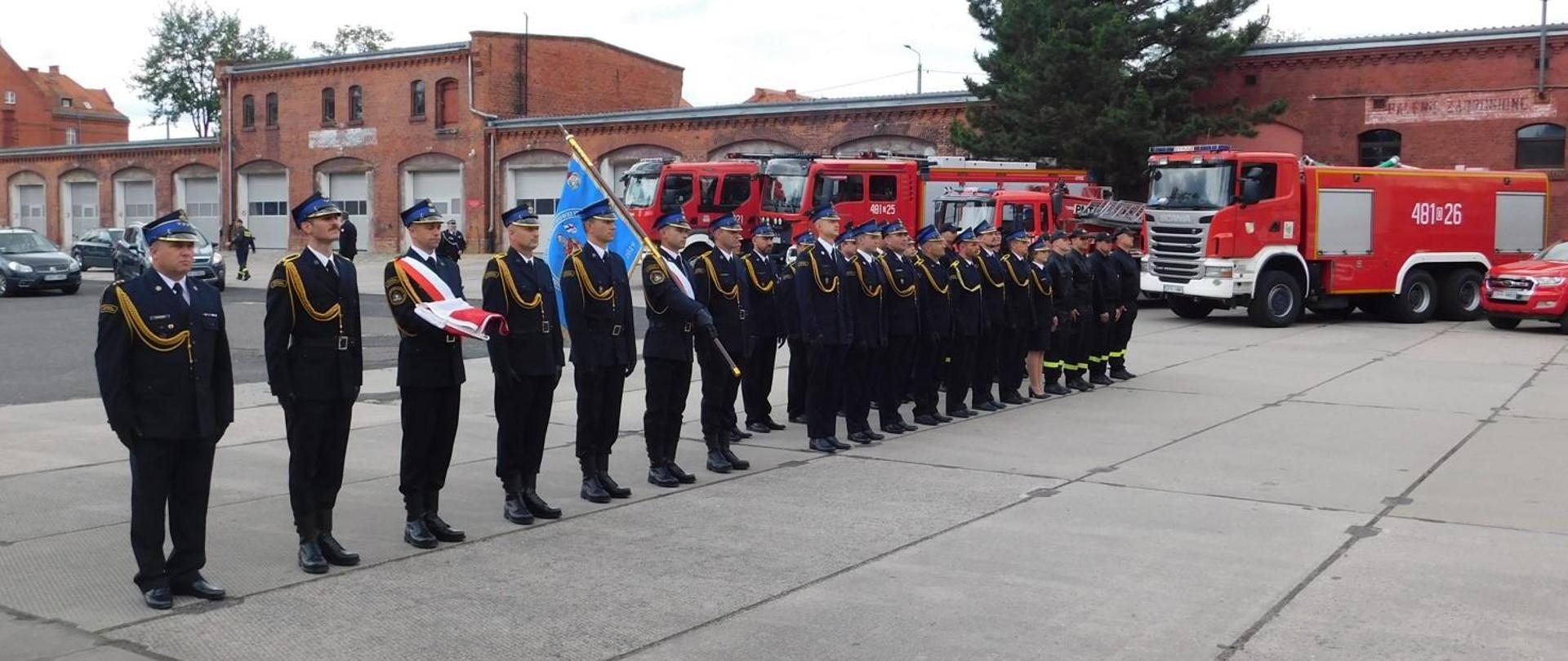 Zdjęcie przedstawia pododdział strażaków na uroczystym apelu z okazji Dnia Strażaka. Na zdjęciu widoczni strażacy w mundurach galowych, poczet flagowy, poczet sztandarowy. W tle pojazdy pożarnicze