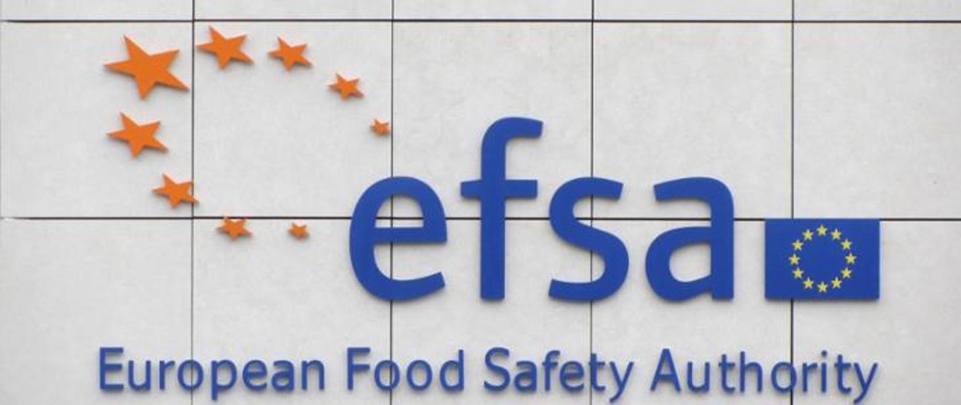 Na zdjęciu znajduje się granatowy napis efsa. Po jego lewej stronie widnieją żółte gwiazdki w okręgu. Po prawej znajduje się flaga Unii Europejskiej. Poniżej widnieje napis European Food Safety Authority. Tło jest w kolorze szarym.