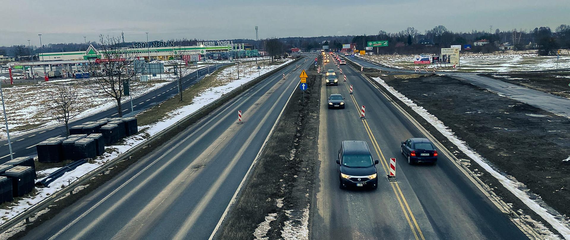 Fotografia przedstawia dwupasmową drogę z ruchem w obu kierunkach. Na pierwszym planie widoczne są samochody jadące lewą jezdnią w obu kierunkach, a na poboczu drogi znajdują się znaki drogowe i pachołki ostrzegawcze, sugerujące prace drogowe lub remont. W tle po lewej stronie widoczny jest duży sklep Leroy Merlin oraz inne budynki handlowe. Na poboczu drogi po lewej stronie widoczne są drzewa oraz pojemniki na odpady. Na poboczu drogi po prawej stronie widoczne są tablice reklamowe oraz pola pokryte śniegiem. Cała scena ma zimowy charakter, z resztkami śniegu na poboczach i trawnikach.