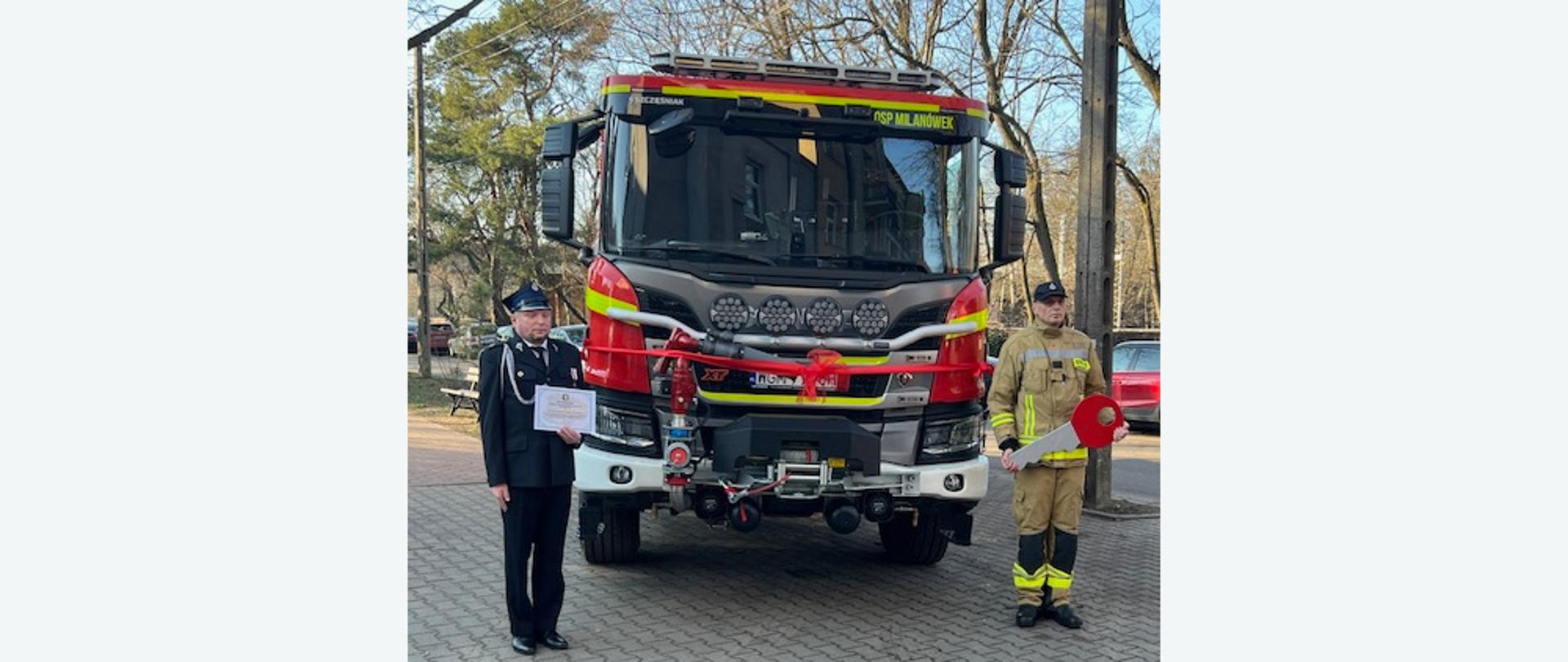 Naczelnik OSP Milanówek stoi z aktem przekazania nowego samochodu pożarniczego, obok stoi kierowca z symbolicznym kluczem do samochodu z tyłu nowy samochód pożarniczy GCBA 10/61 SCANIA