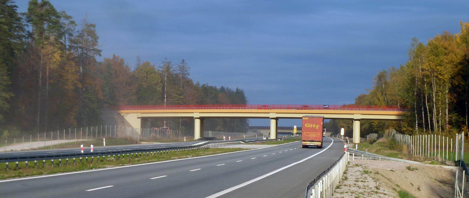 S7 obwodnica Kielc - wiadukt pomalowany na czerwono i pomarańczowo nad dwujezdniową drogą ekspresową. W oddali przejście dla zwierząt nad drogą. Dwie ciężarówki jadące drogą. Po bokach drogi bariery, siatki ochronne. Za nimi drzewa.