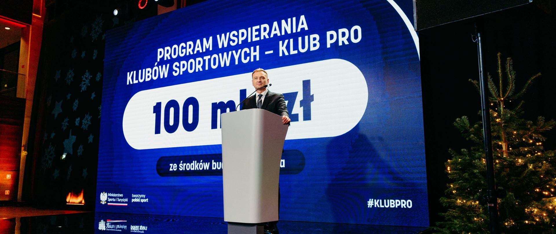 Sławomir Nitras stoi na scenie za biała mównicą. Za nim granatowy slajd z napisem: "Program wspierania klubów sportowych - KLUB PRO. 100 mln zł ze środków budżetu państwa. Na dole w lewym roku logotypy MSiT i napis tworzymy polski sport. W prawym dolnym rogu hashtag #KLUB PRO. Z prawej strony zdjęcia choinka z lampkami.