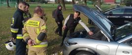 Teren stadionu w Rawiczu. Instruktor z salonu sprzedaży pojazdów prezentuje auto elektryczne. Obok niego uczestnicy warsztatów w ubraniach specjalnych. W tle boiska i drzewa.