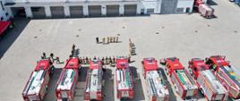 Zdjęcie zrobione z wysokości. Na placu wewnętrznym jednostki ratowniczo- gaśniczej stoją pojazdy pożarnicze. przed nimi stoją zgromadzeni funkcjonariusze. W tle widać strażackie bramy garażowe i budynki koszar wojskowych. 