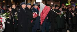 Plac przed Pomnikiem Żołnierza Polskiego w Rawiczu, na nim stoją uczestnicy uroczystego apelu. Wśród nich jest delegacja rawickich służb mundurowych. W środku stoi strażak PSP trzymający wiązankę kwiatów. Obok mężczyzna z biało-czerwona flagą. 