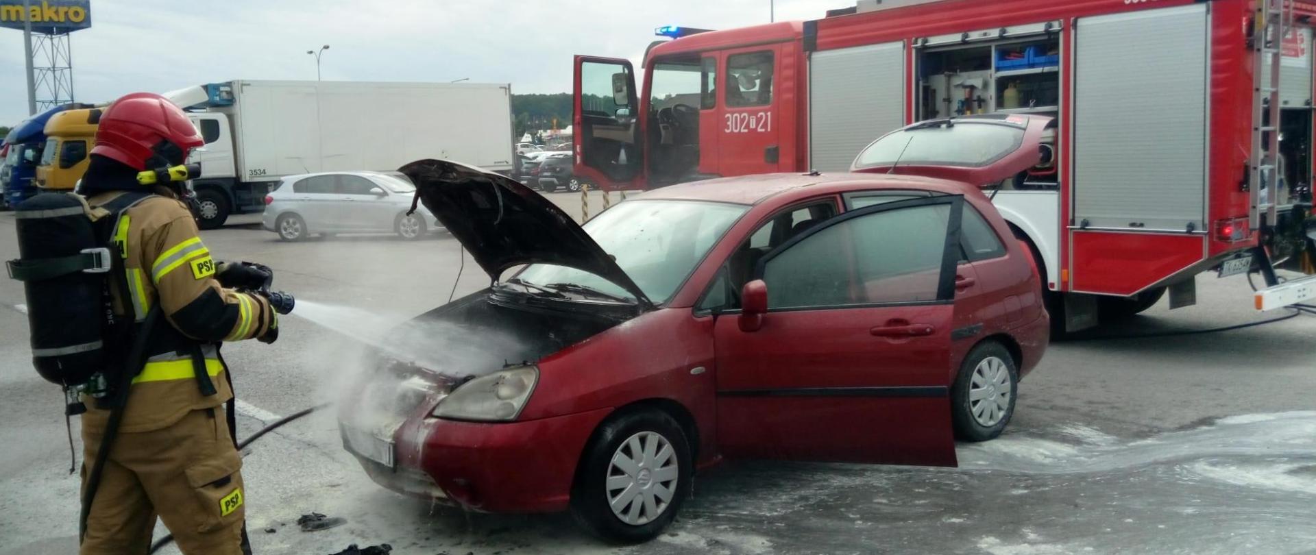 Zdjęcie przedstawia samochód, który uległ spaleniu. Przed nim ratownik podający pianę na komorę silnika, a z tyłu widać zaparkowany samochód pożarniczy.
