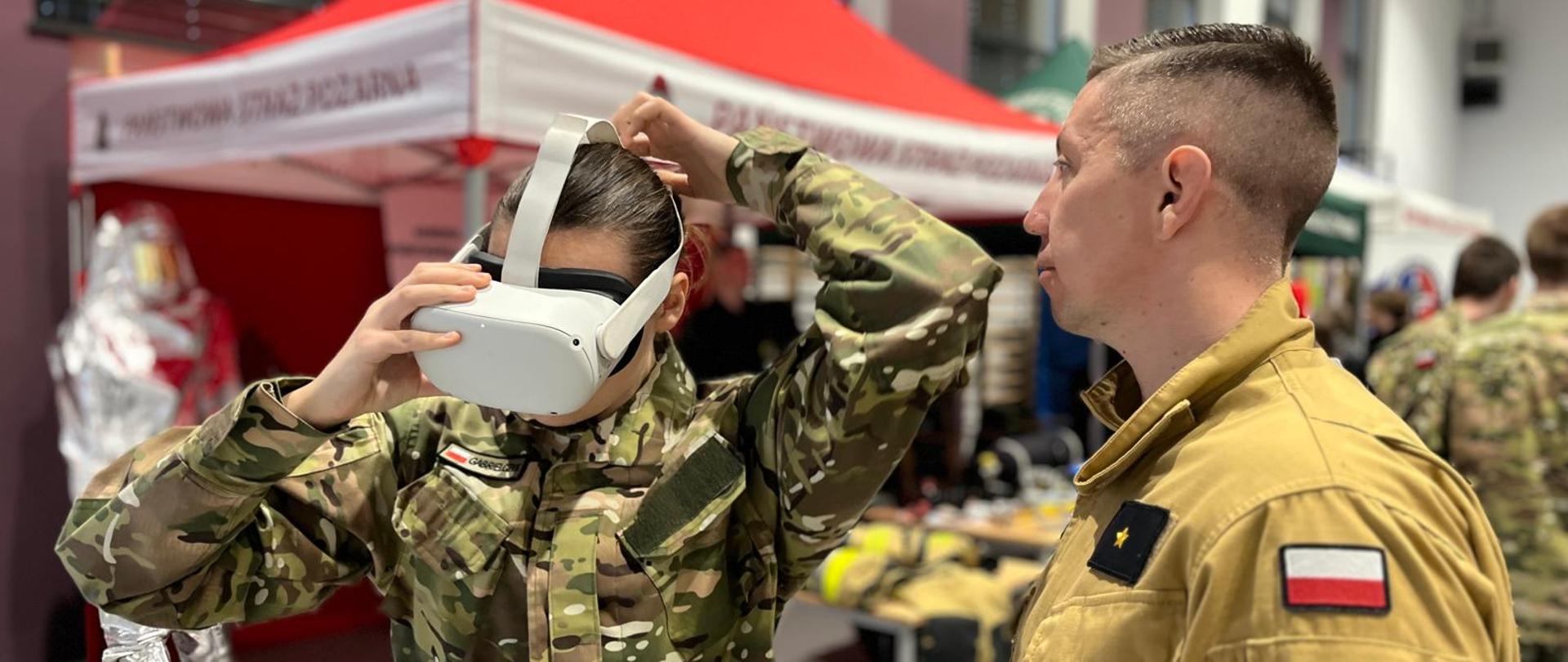 Strażak w mundurze służbowym pomaga dziewczynie założyć gogle VR dla celów prezentacji. Na rękawie strażaka widoczne logo PSP i flaga narodowa biało-czerwona. W tle namiot reklamowy, czerwony. 