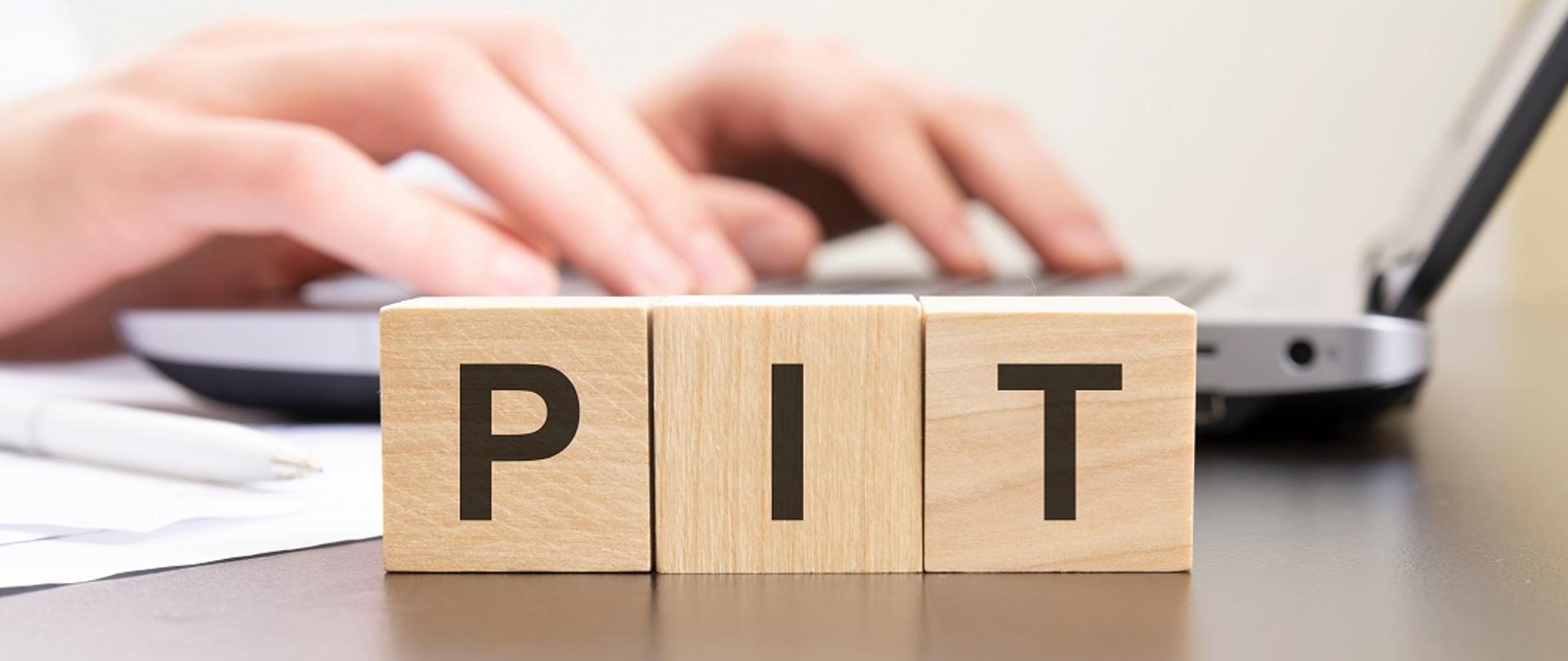 Akronim PIT na drewnianych klockach. W tle dłonie na laptopie.
pit - acronym from wooden blocks with letters. background hands 