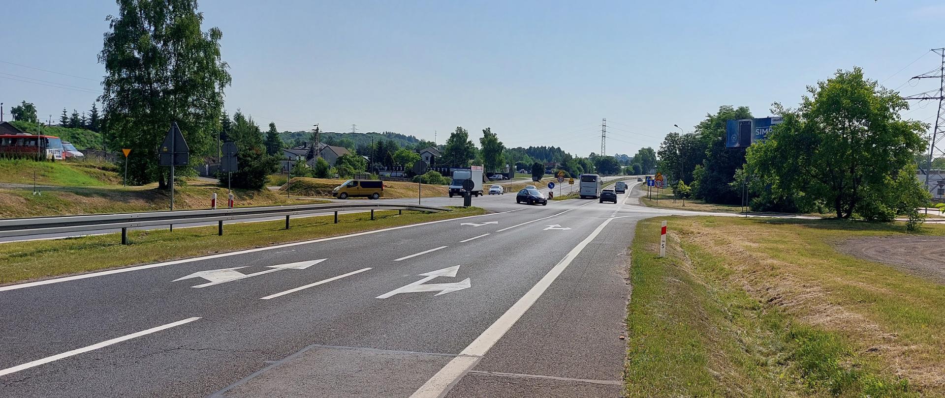 Zdjęcie drogi krajowej nr 94 w Olkuszu. Widoczne dwie jezdnie po dwa pasy ruchu w obu kierunkach z oznakowaniem poziomym i pionowym. Jezdnie oddzielone są pasem zieleni i barierami ochronnymi. 