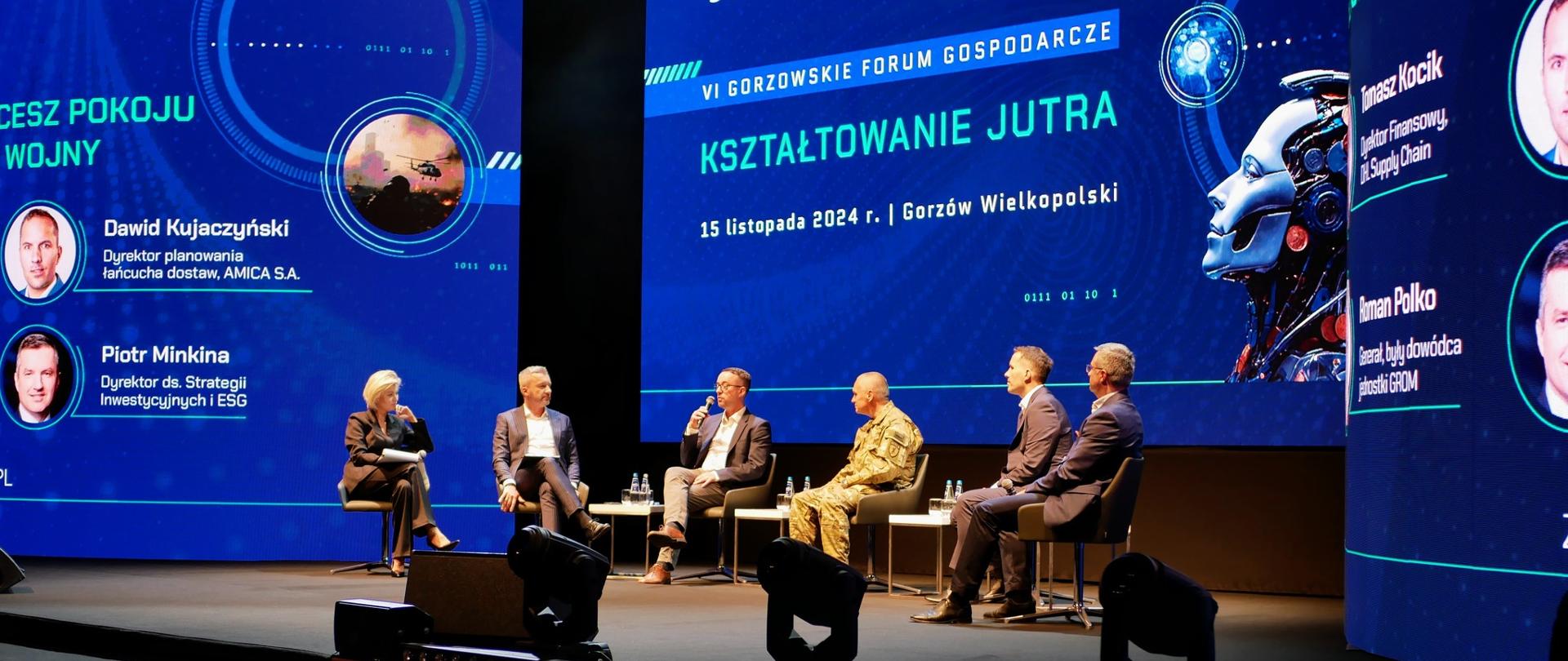 Widok na scenę na której siedzi 6 osób prowadzących panel dyskusyjny. W tle Niebieskie banery reklamowe