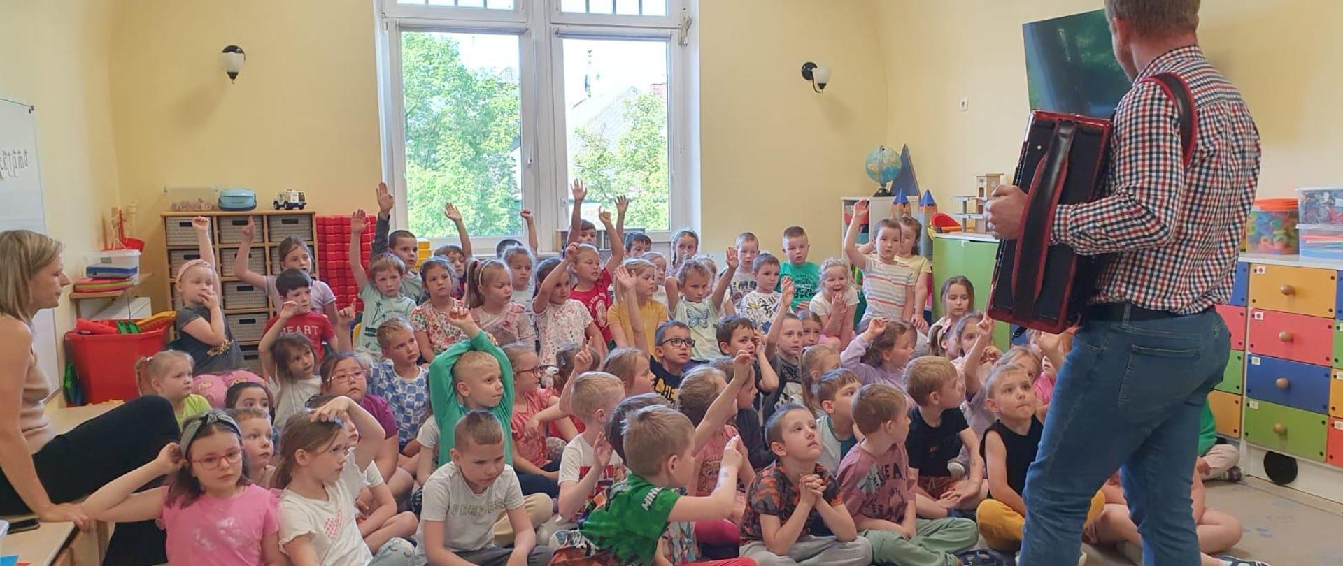 Grupa dzieci siedzących na podłodze w przedszkolu, a na akordeonie gra nauczyciel Tomasz Brusiński w Przedszkolu nr 1 w Kłodzku.