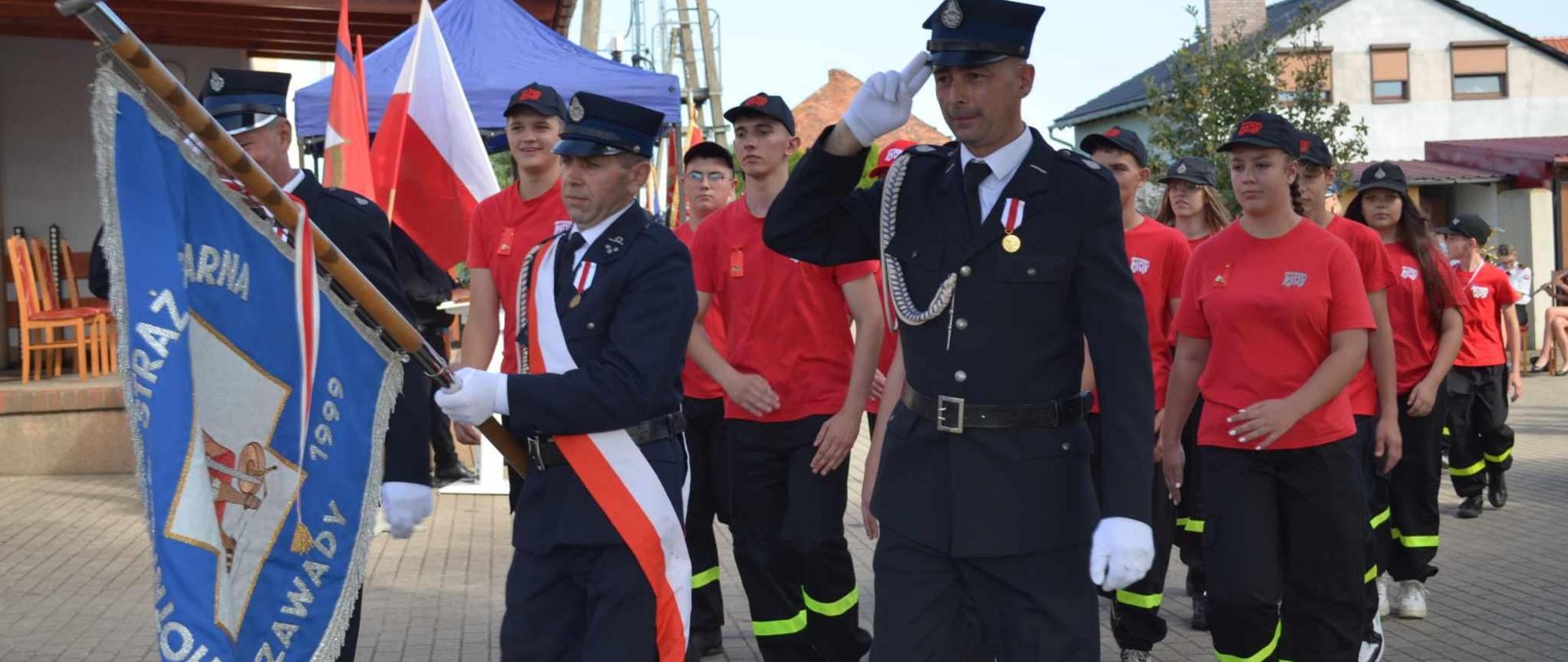 Plac przed świetlicą wiejską, na którym odbywa się uroczysty apel. Odbywa się defilada pododdziałów. Na pierwszym planie poczet sztandarowy OSP w Zawadach, za nim członkowie MDP w czerwonych koszulkach i czapkach. W tle budynki.