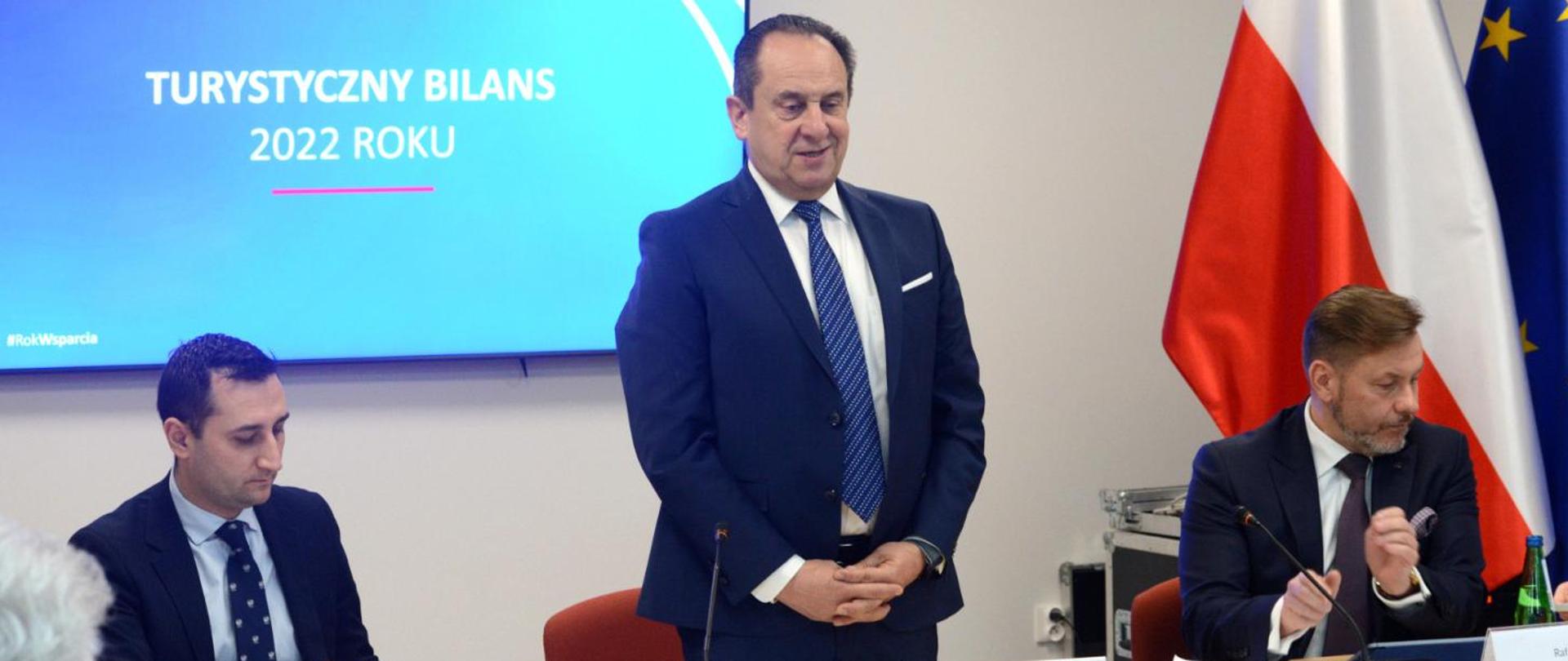 three men in suits, minister is standing in front of the desk, there is a screen behind him