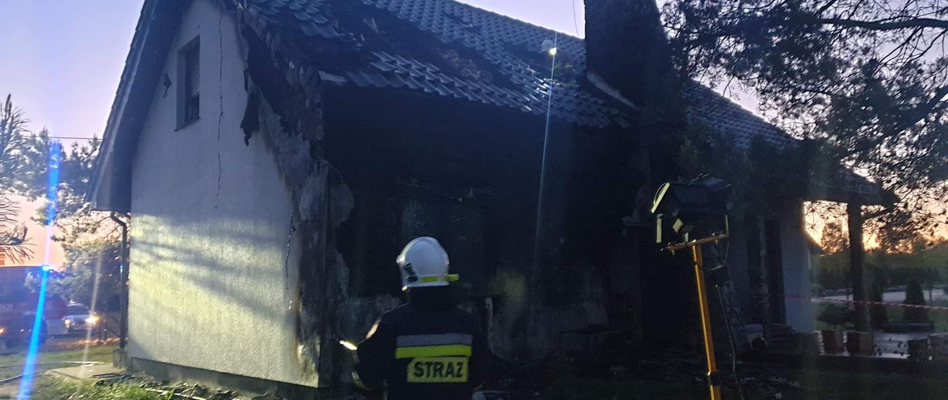 Zdjęcie przedstawia budynek mieszkalny jednorodzinny, w którym miał miejsce pożar. Dach budynku częściowo zniszczony oraz elewacja frontowa również. Na zdjęciu widać również strażaka