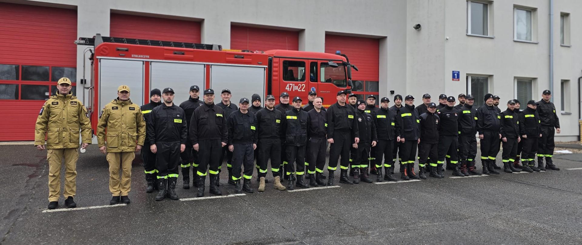 Grupa strażaków w ubraniach koszarowych koloru czarnego. Dwóch funkcjonariuszy w ubraniach służbowych koloru piaskowego na tle samochodu bojowego i budynku komendy powiatowej