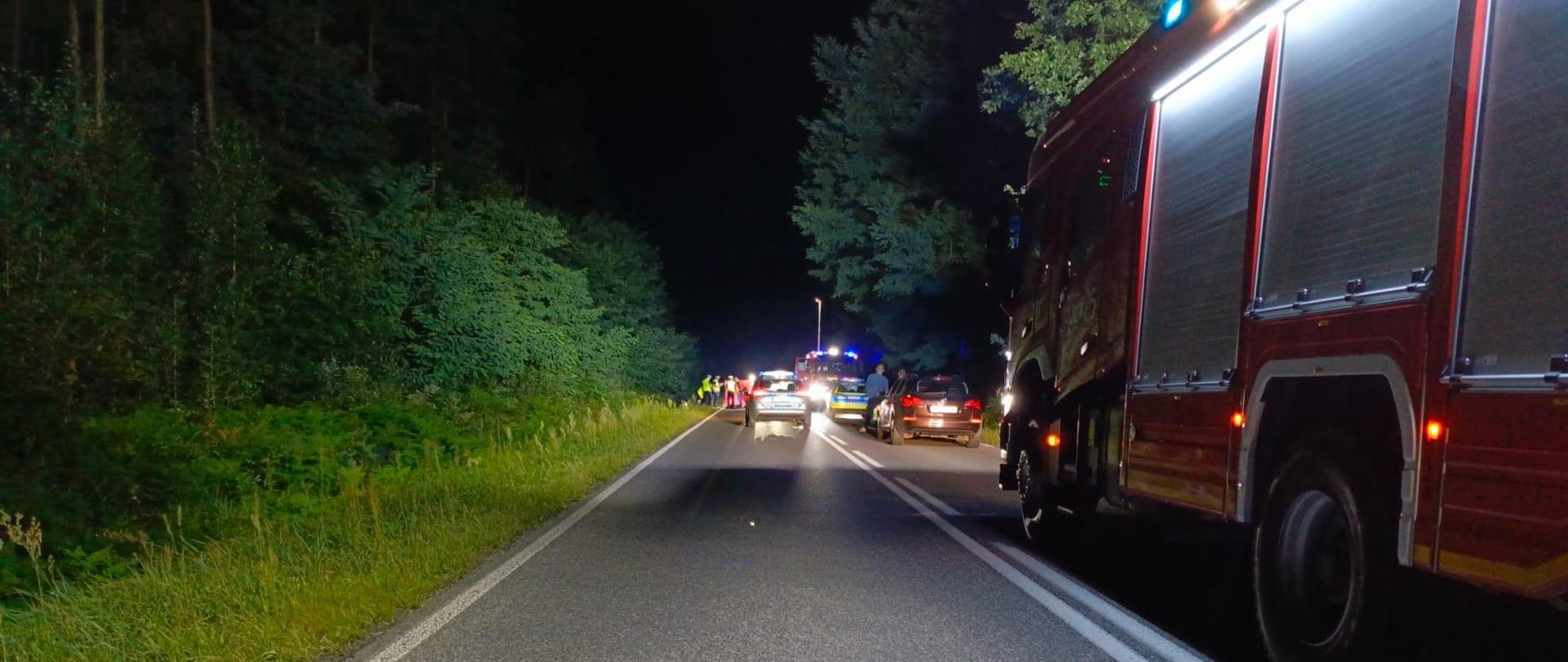 Zdjęcie przedstawia drogę wojewódzką – widok z góry, na której stoją pojazdy służb ratowniczych. Głęboko w oddali, po lewej stronie, czerwony parawan osłania miejsce udzielania pomocy poszkodowanemu. 