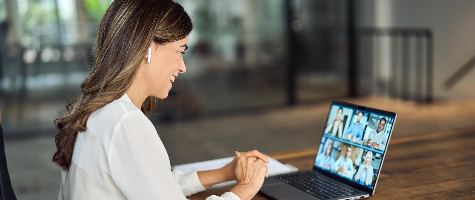 Ujęcie boczne, kobieta siedząca przy biurku. Na blacie laptop, kartki i telefon. Na ekranie widoczni uczestnicy spotkania online.