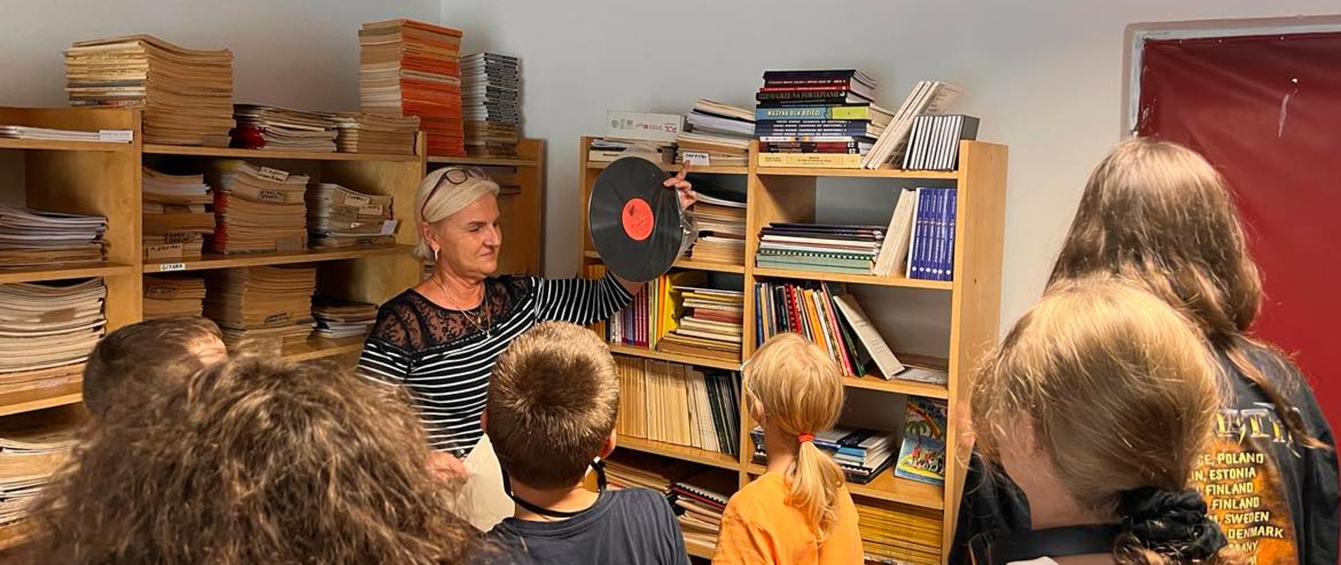 Zdjęcie przedstawiające uczniów klasy I A i III cyklu 4-letniego w bibliotece szkolnej. W tle nauczyciel-bibliotekarz - mgr Iwona Florkowska - pokazujący płytę winylową.