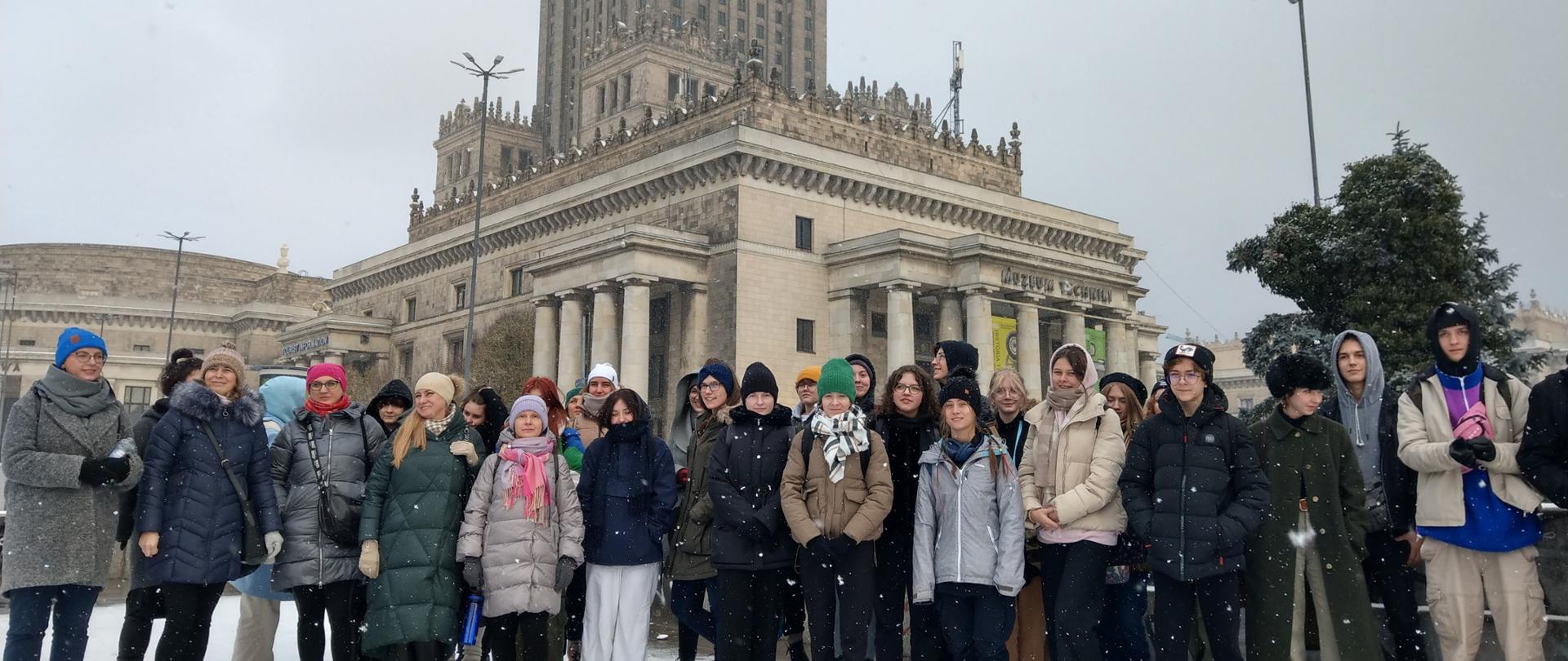 Zdjęcie przedstawia kilkudziesięcioosobową grupę młodzieży i wychowawców, wszyscy ubrani w zimowe kurtki czapki szaliki rękawice w różnych kolorach. Stoją na tle Pałacu Kultury i Nauki W Warszawie. Widać padajace płatki śniegu.