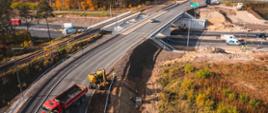 Obraz przedstawia plac budowy drogi lub autostrady. Na pierwszym planie widać wiadukt, po którym poruszają się pojazdy. Pod wiaduktem trwają prace budowlane. Na drodze widoczne są czerwony wywrotka oraz żółta koparka, która pracuje na poboczu. Widać również kilku pracowników budowlanych i materiały konstrukcyjne. Otoczenie obejmuje tory kolejowe, drzewa w jesiennych barwach oraz inne pojazdy i sprzęt budowlany. Zdjęcie oddaje złożoność i skalę nowoczesnych prac infrastrukturalnych, ukazując współpracę różnych elementów, takich jak pojazdy, maszyny i pracownicy.