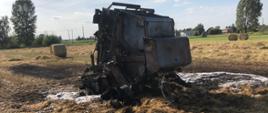 Zdjęcie przedstawia spaloną prasę rolniczą na polu uprawnym. W tle sprasowana słoma w balotach oraz rżysko. 