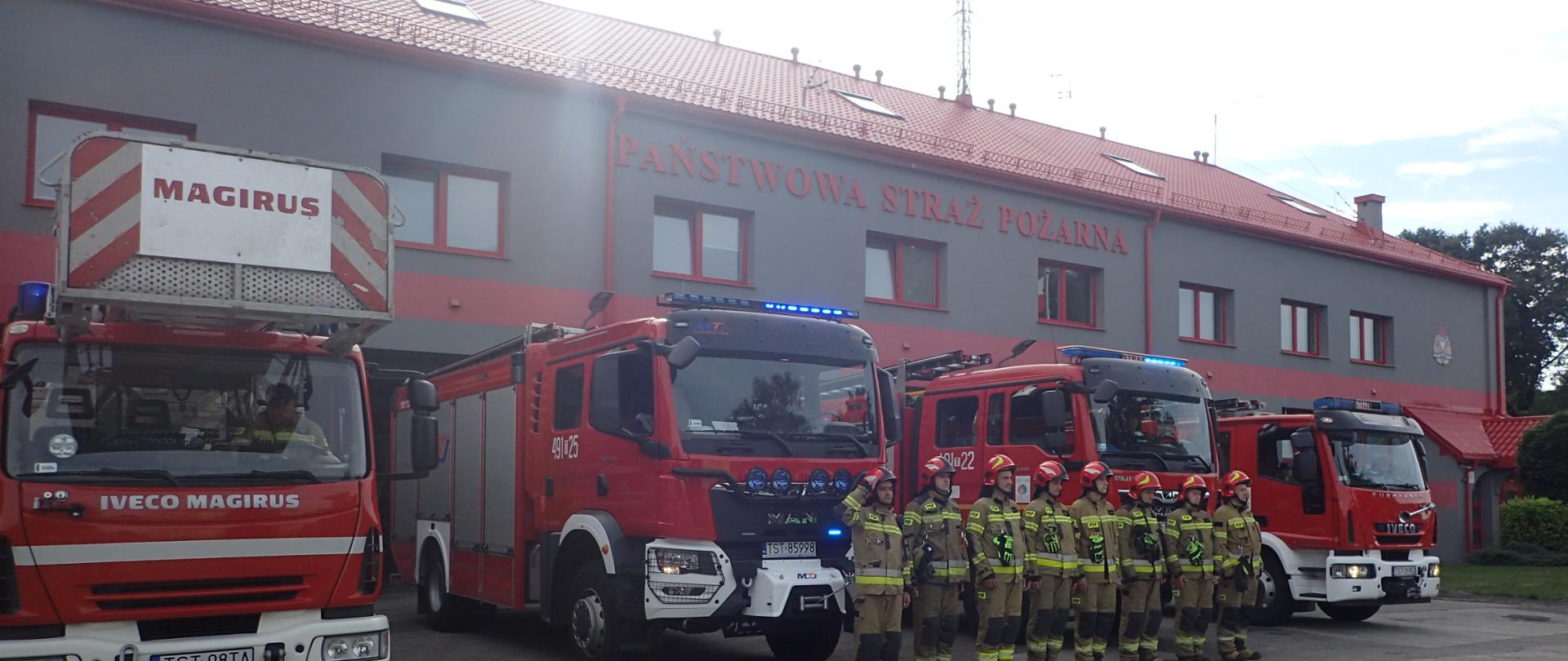 Strażacy oddający honory bohaterom powstania warszawskiego, w tle wozy bojowe oraz budynek KP PSP