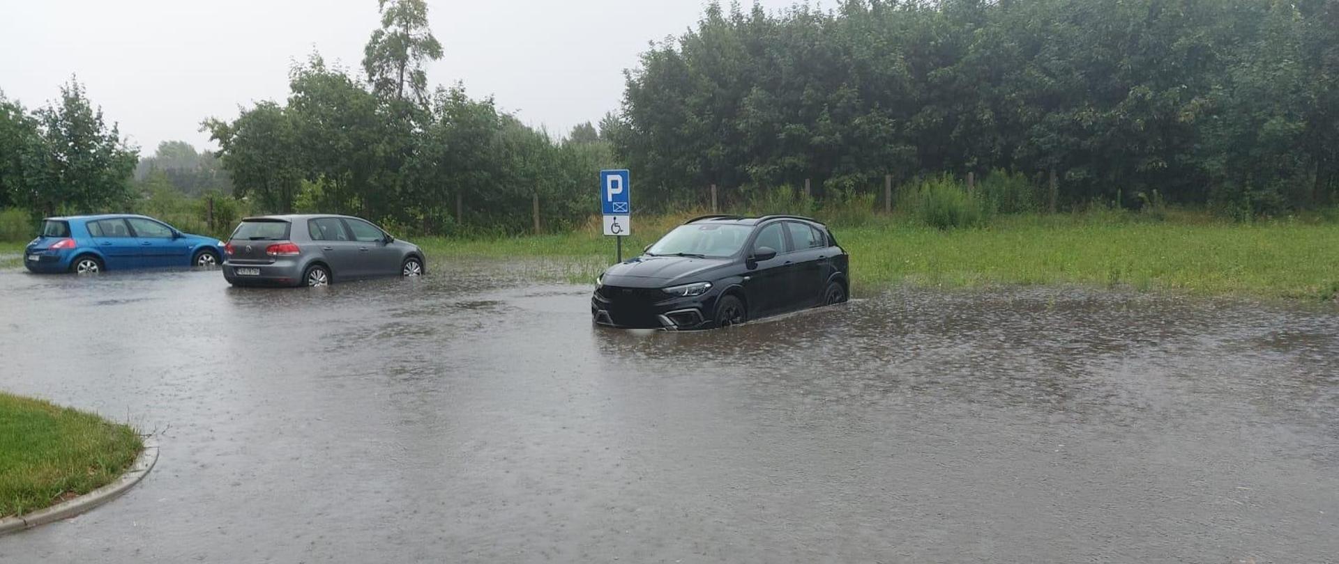 Zdjęcie przedstawia trzy samochody osobowe stojące na parkingu, które zostały podtopione przez opady deszczu.
