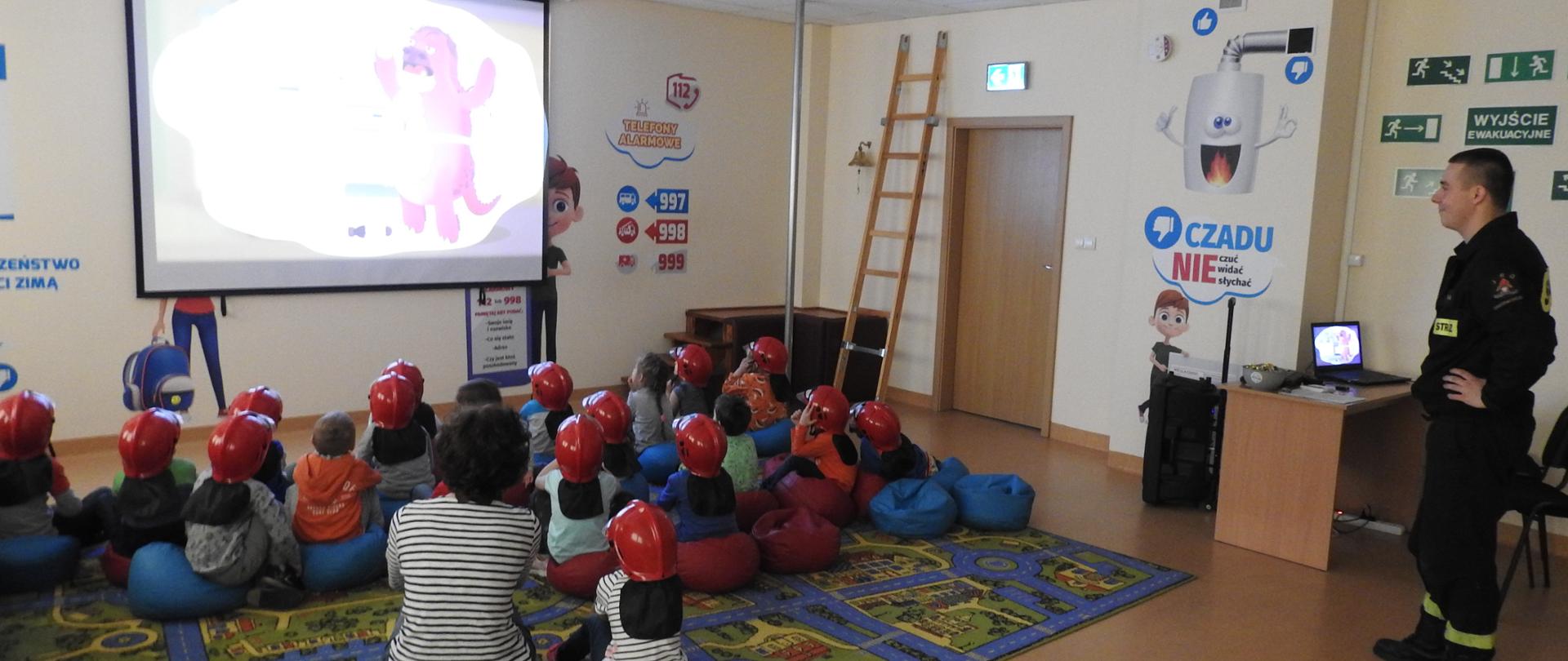 Dzieci siedzą na kolorowym dywanie. Oglądają film. Na głowach mają czerwone hełmy. Za nimi obok biurka stoi strażak. Strażak też ogląda film. 
