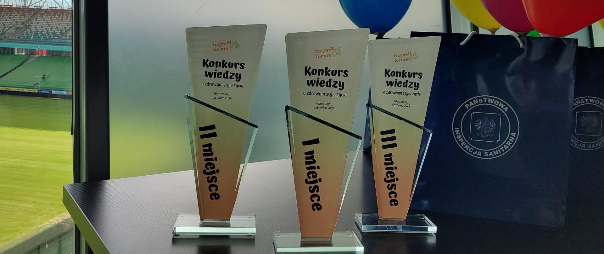 Trzy szklane statuetki - nagrody w konkursie "Trzymaj Formę!". W tle stadion Legii.