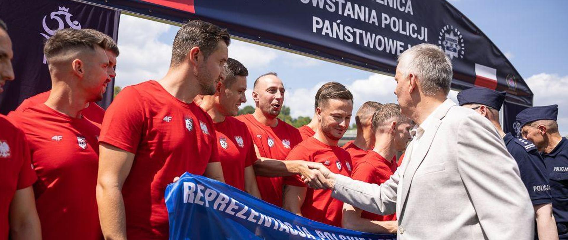 Minister Tomasz Siemoniak wita się z piłkarzami reprezentacji Polskiej Policji poprzez podanie im dłoni. Piłkarze mają czerwone stroje piłkarskie, trzymają niebieską flagę reprezentacji Polskiej Policji.