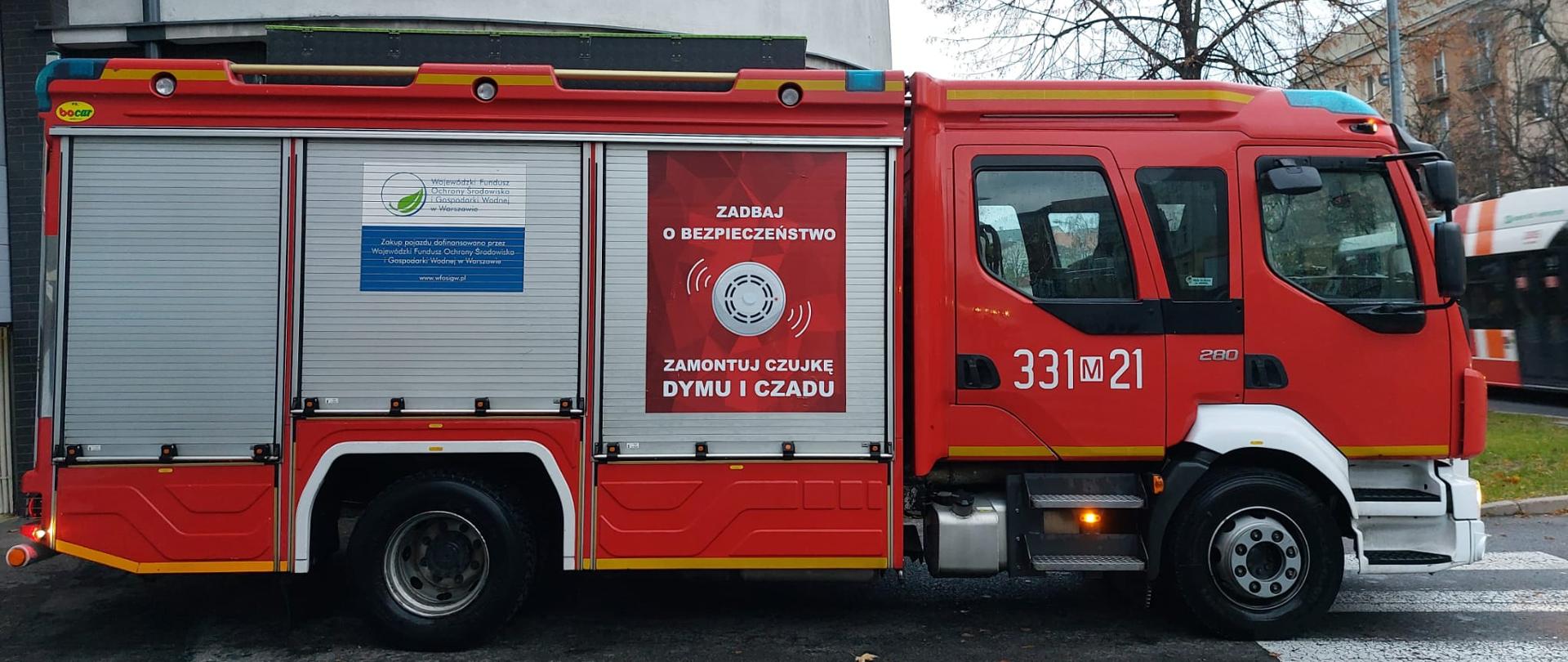 Na zdjęciu widać pojazd pożarniczy, na którym od prawej strony na pierwszej rolecie skrytki naklejony jest plakat "Zadbaj o bezpieczeństwo Zamontuj czujkę dymu i czadu".