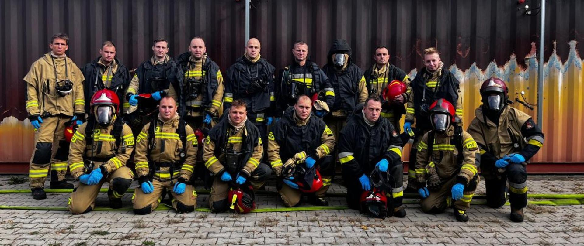 Wspólne zdjęcie ćwiczących oraz instruktorów na tle wielofunkcyjnego trenażera ratowniczego. Zdjęcie przedstawia strażaków Państwowej Straży Pożarnej w umundurowaniu bojowym na poligonie ratowniczym Komendy Wojewódzkiej Państwowej Straży Pożarnej.