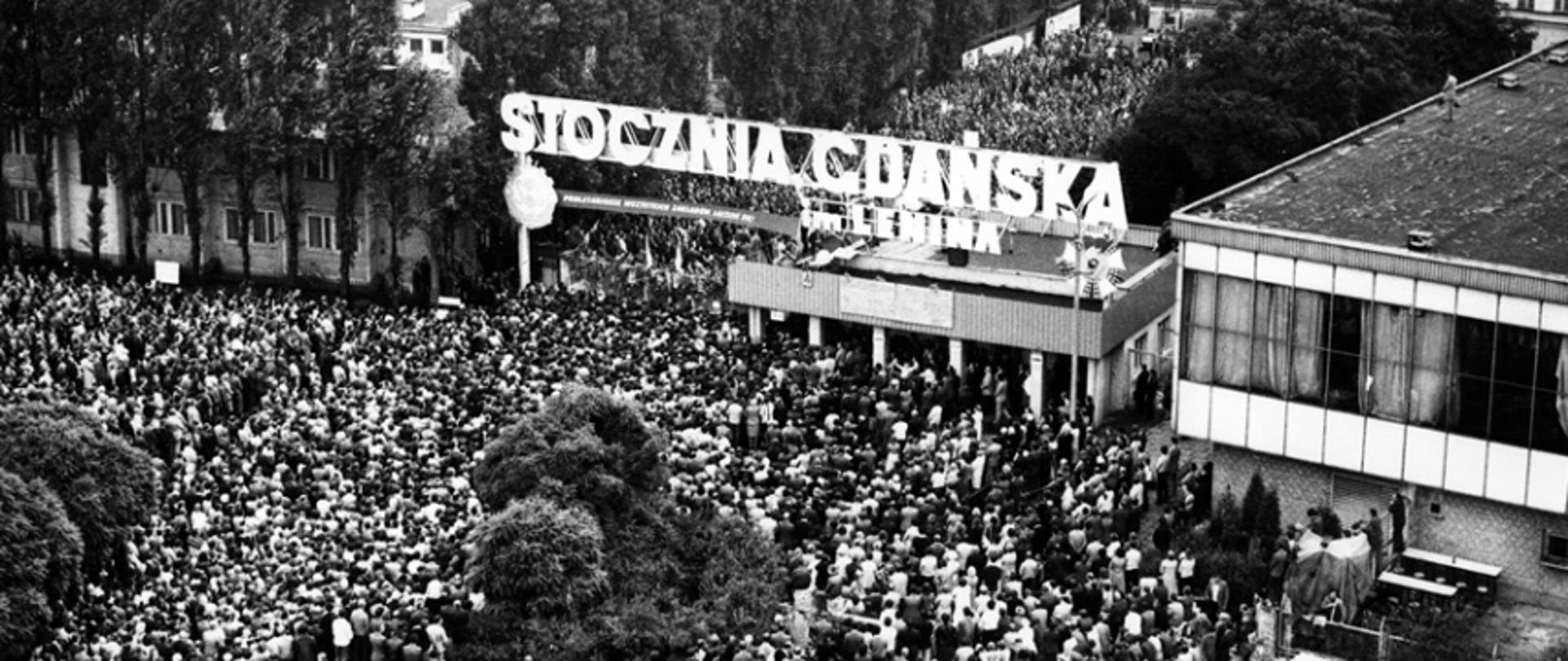 August strike at Lenin's Shipyard in Gdansk. Top view of Gate No. 2 and the citizens and shipyard workers gathered below it. Above the gate, there is a banner with the slogan "PROLETARIANS OF ALL FACTORIES UNITE!", and on the roof of the guardhouse there are boards with 21 demands of MKS.
