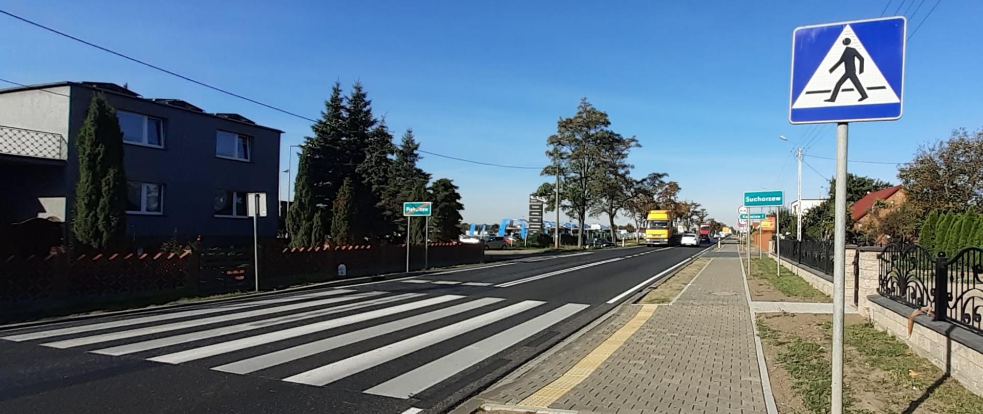 Zdjęcie przedstawia drogę krajową nr 11 w miejscowości Piekarzew. Na zdjęciu widoczna droga, po jednym pasie w każdym kierunku. Z prawej strony chodnik wraz z przejściem dla pieszych. W oddali widoczne samochody.