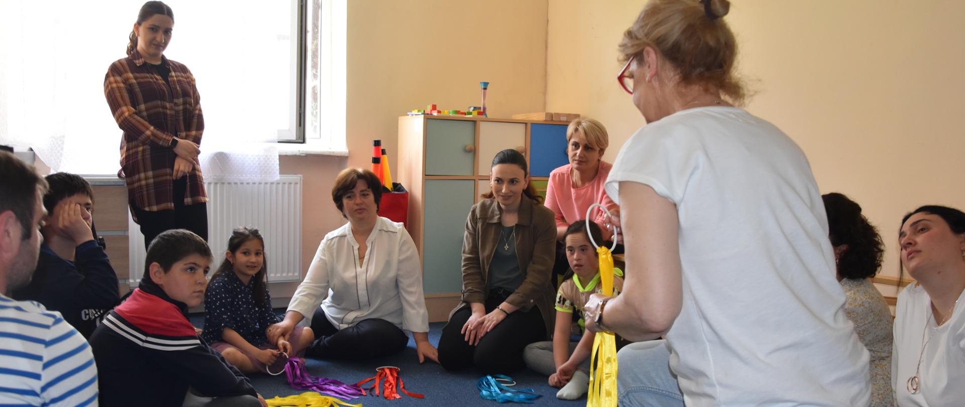 Na podłodze w półkolu siedzą dorośli z dziećmi i patrzą się na instruktorkę, która trzyma w ręku kółko z taśmami; podobne kółka z taśmami leżą na podłodze.