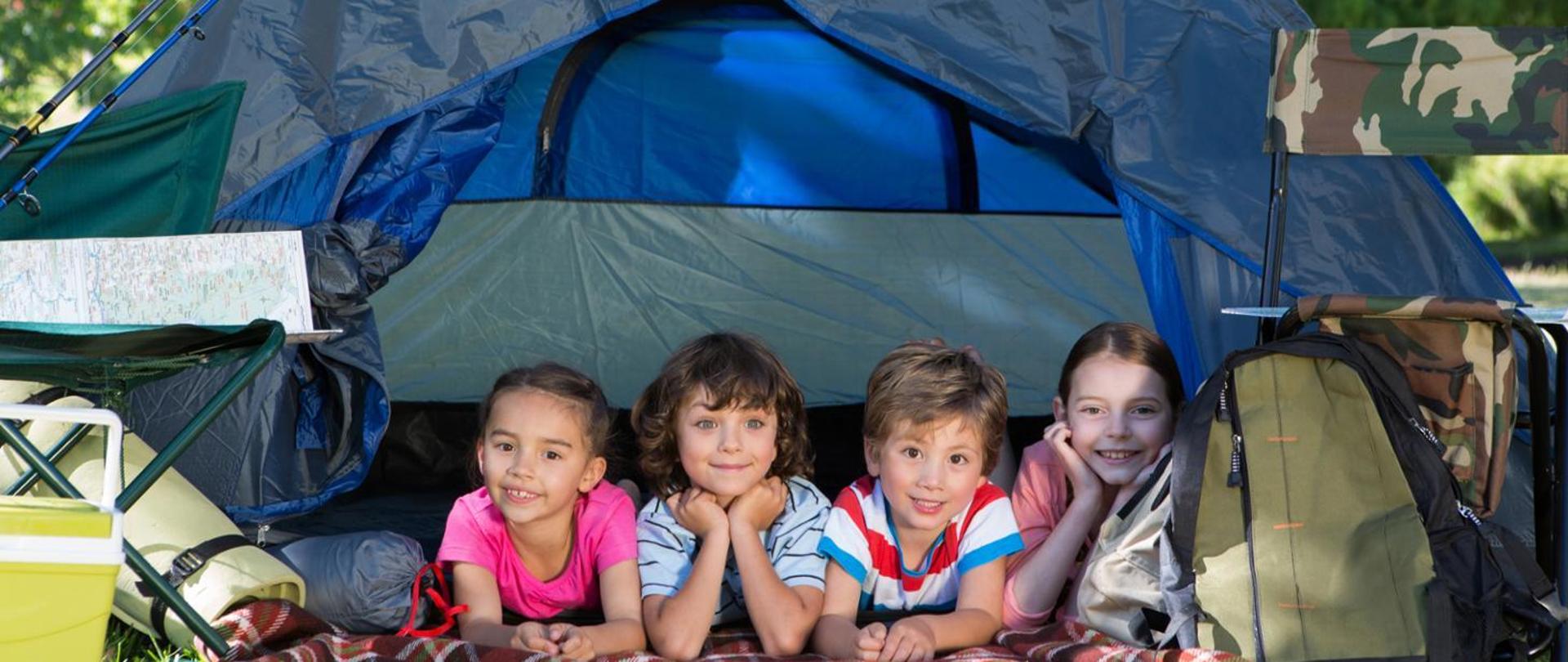 czworo dzieci wypoczywających pod namiotem obok stoją krzesełka turystyczne, plecak, śpiwór, mapa