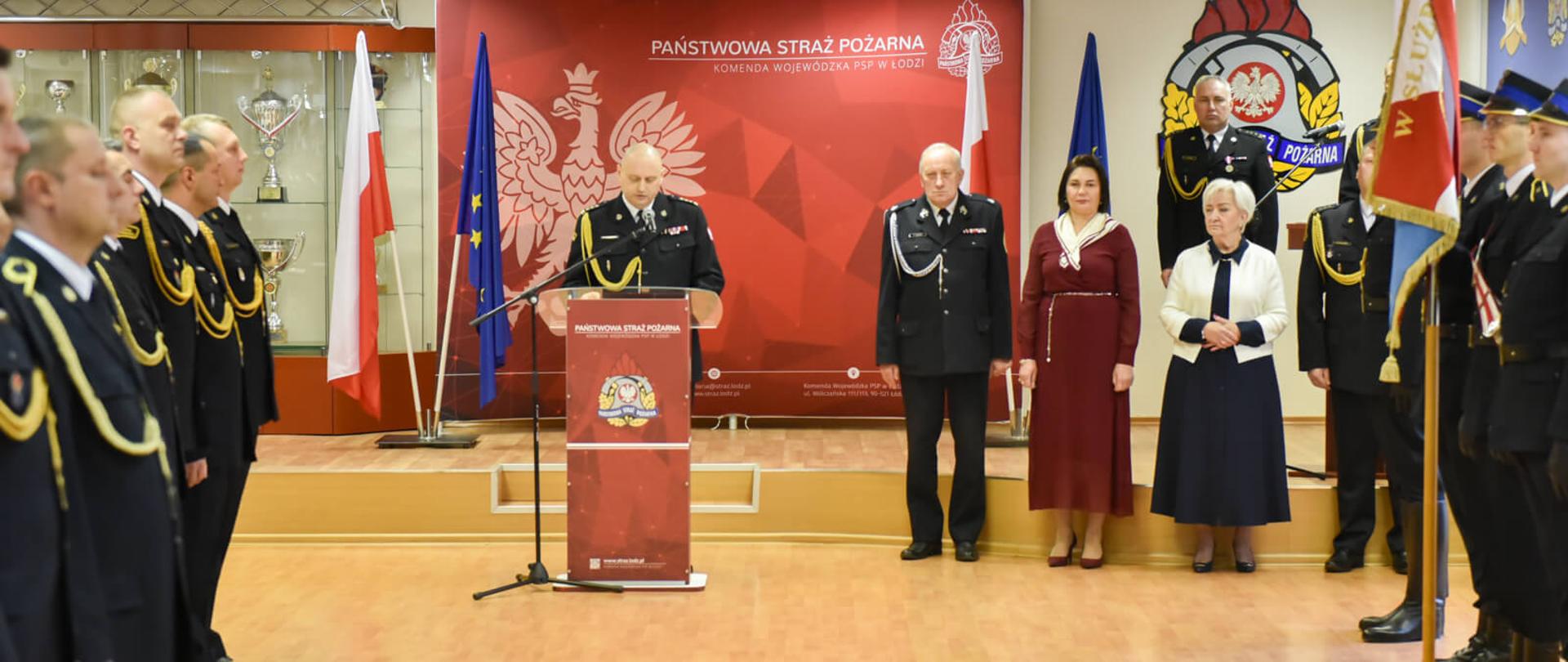 strażacy i uczestnicy wydarzenia stoją na zbiórce w sali konferencyjnej, po lewej stoją strażacy w szeregu, na środku stoją komendanci i zaproszeni goście, po prawej poczet sztandarowy