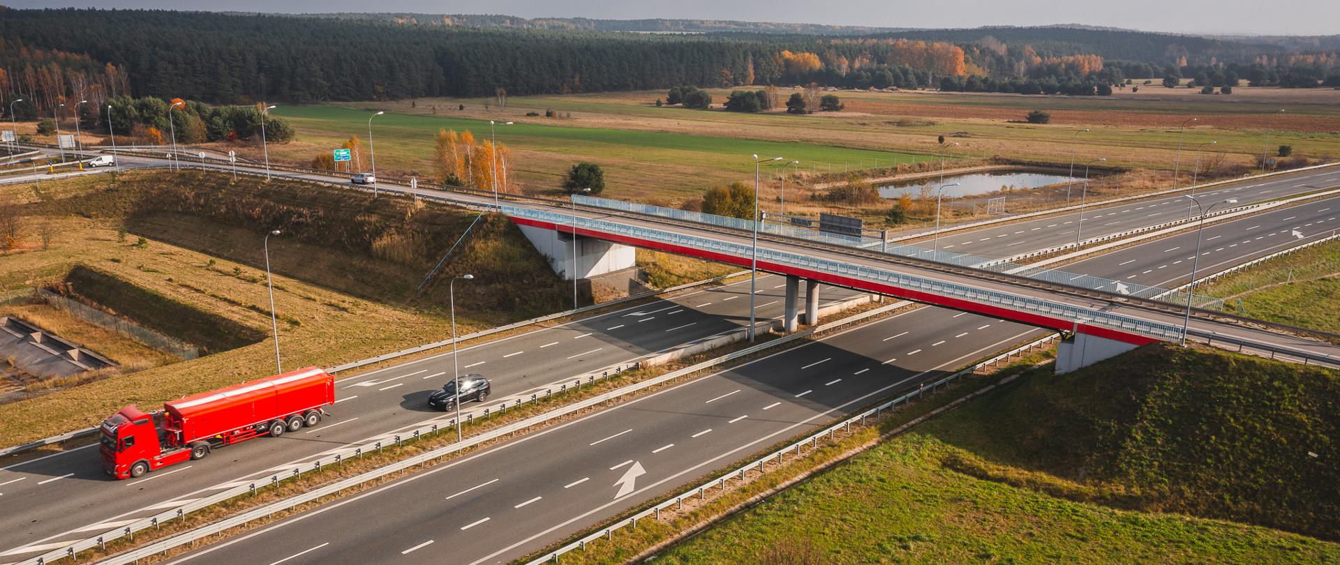 Zdjęcie przedstawia widok z lotu ptaka na autostradę z wiaduktem. Na autostradzie widać czerwony samochód ciężarowy oraz czarny samochód osobowy. Wiadukt przebiega nad autostradą, a w tle widać pola, lasy i kilka drzew. Na autostradzie są trzy pasy ruchu w każdym kierunku, oddzielone barierkami. Wiadukt jest pomalowany na biało i czerwono. Cała scena jest oświetlona naturalnym światłem dziennym, co sugeruje, że zdjęcie zostało zrobione w ciągu dnia.