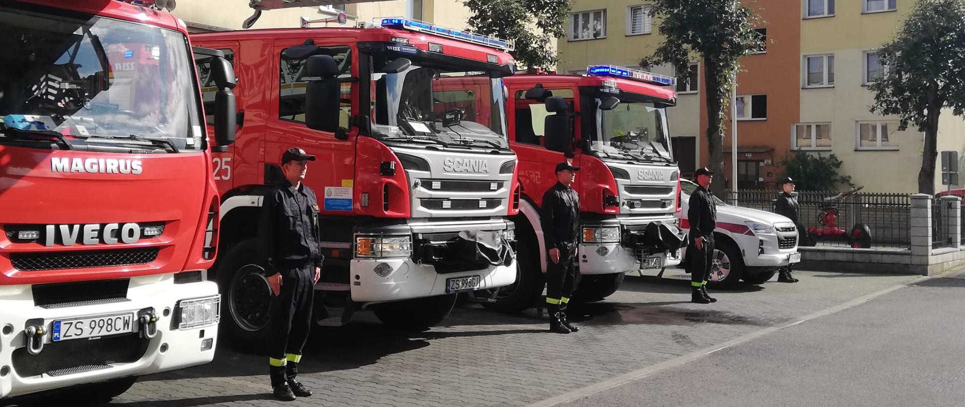 Zdjęcie przedstawia strażaków stojących w postawie zasadniczej pomiędzy samochodami pożarniczymi koloru czerwonego. Strażacy w umundurowaniu koszarowym koloru czarnego.