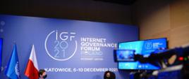 Szczyt Cyfrowy ONZ – IGF 2021. Mężczyzna i kobieta rozmawiający przed kamerą. Z tyłu logo IGF i ekran. Po lewej stronie flagi ONZ i Polski. Fot. PAP